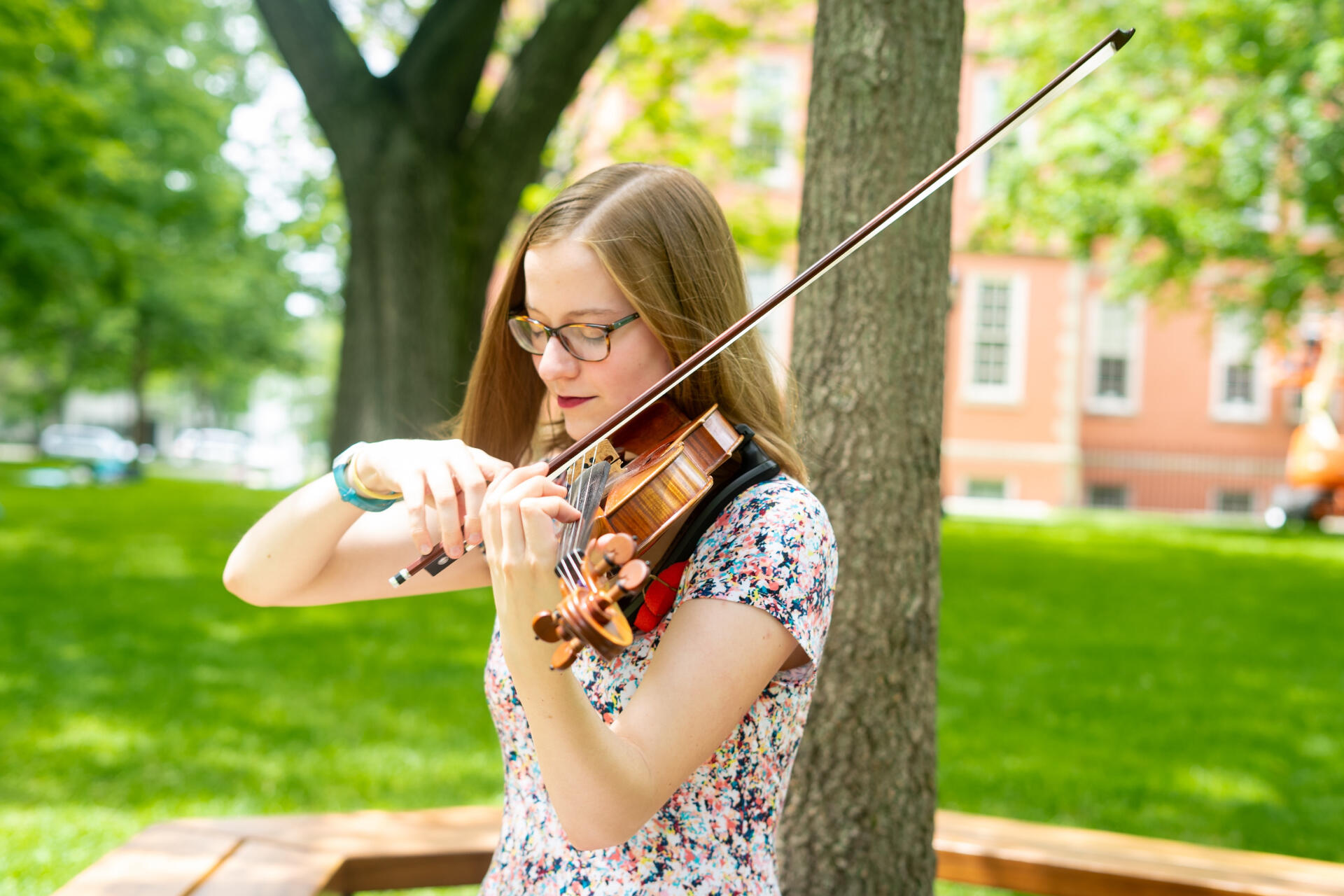 Landscape-mode photograph of Sabine playing her violin next to a tree trunk outside. Her eyes are closed as she plays.