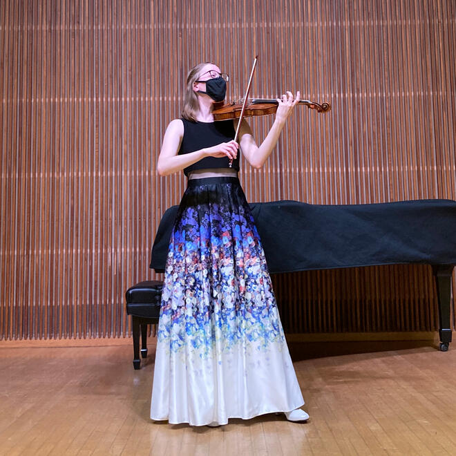 Full-length photograph of Sabine playing her violin in a recital hall. She is wearing a full length dress.
